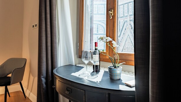 A small room with a window, a chair, a table with a bottle of wine, two wine glasses, and a potted orchid. - Florence Dome Rooms (Florence)