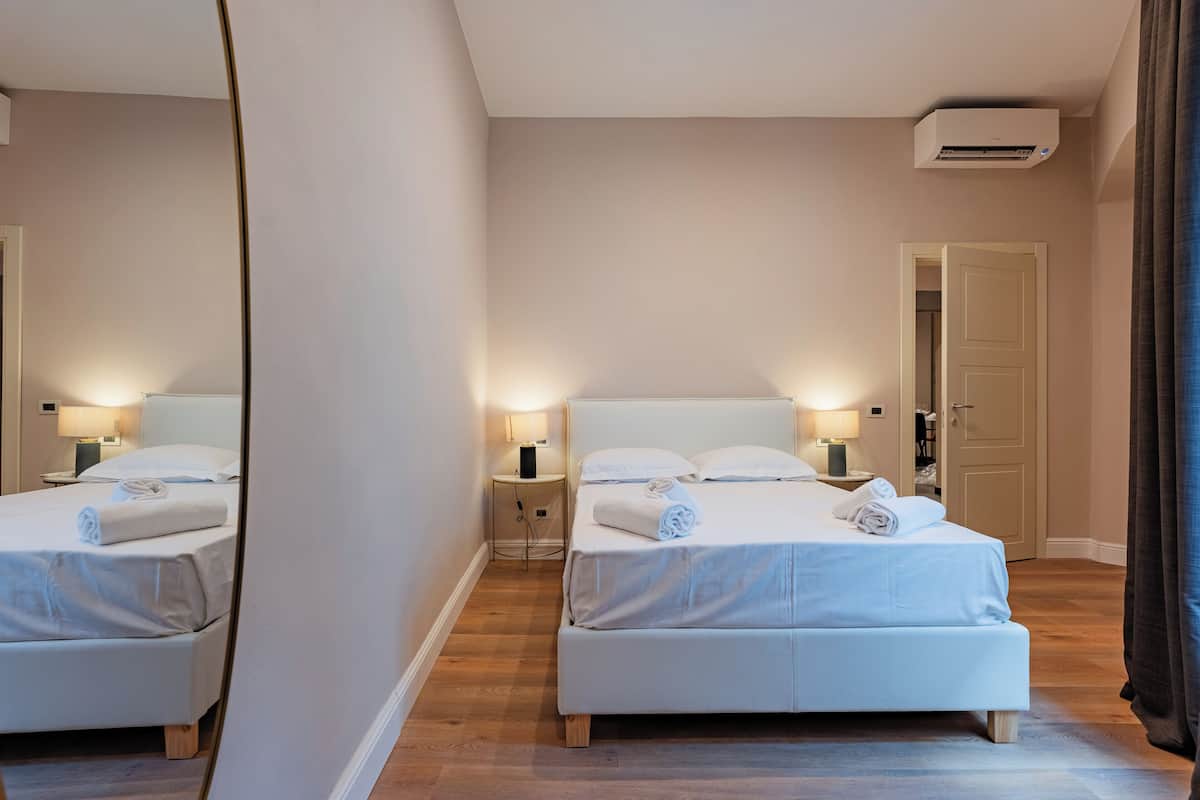 A hotel room with two beds, bedside tables with lamps, an air conditioning unit, and a door.