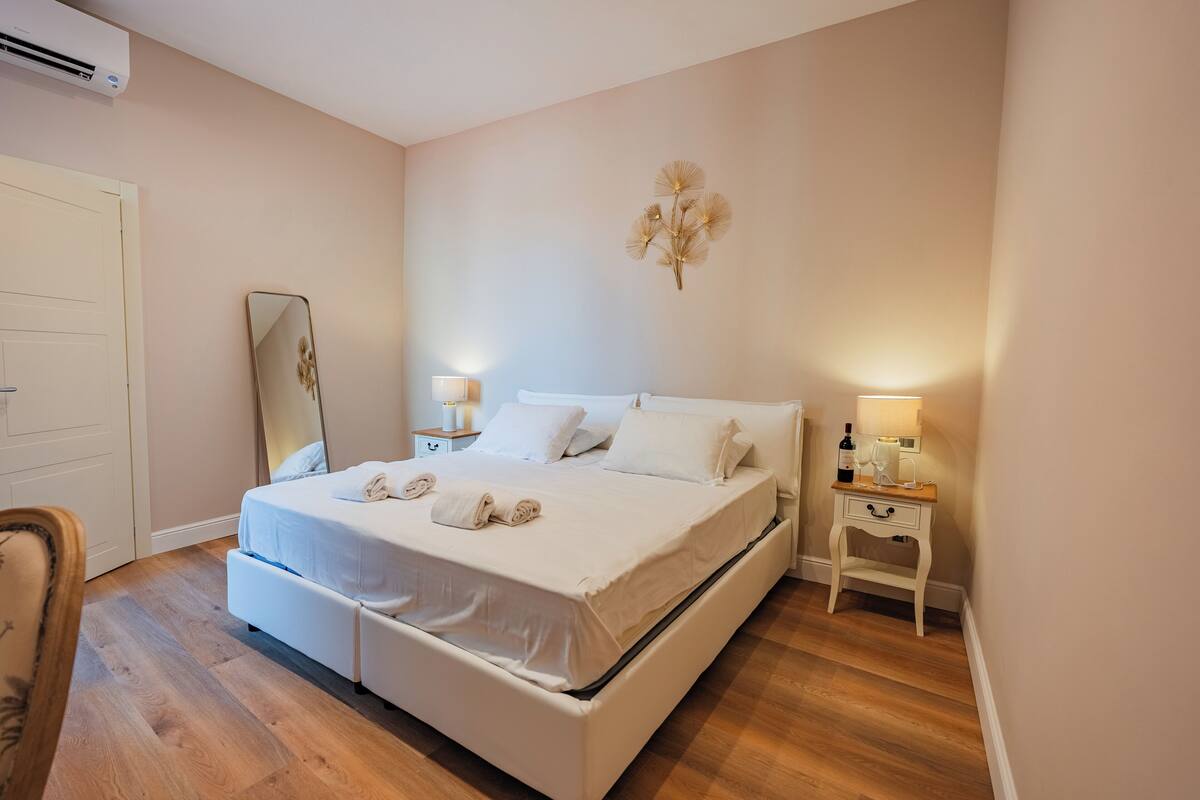 A neatly made bed with white linens, a bedside table with a lamp, and a wall-mounted mirror.