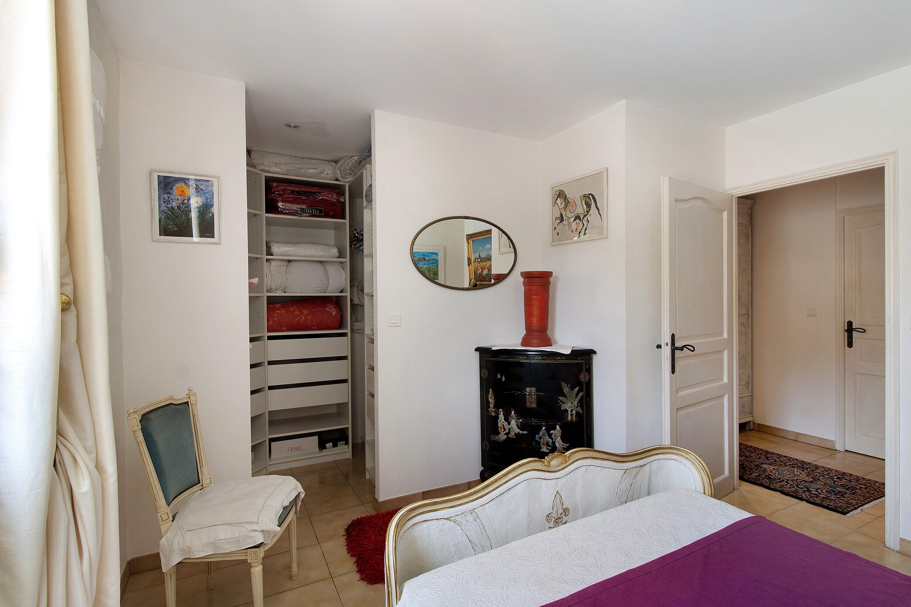 A bedroom with a bed, a chair, a wardrobe, a mirror, and a door leading to another room.