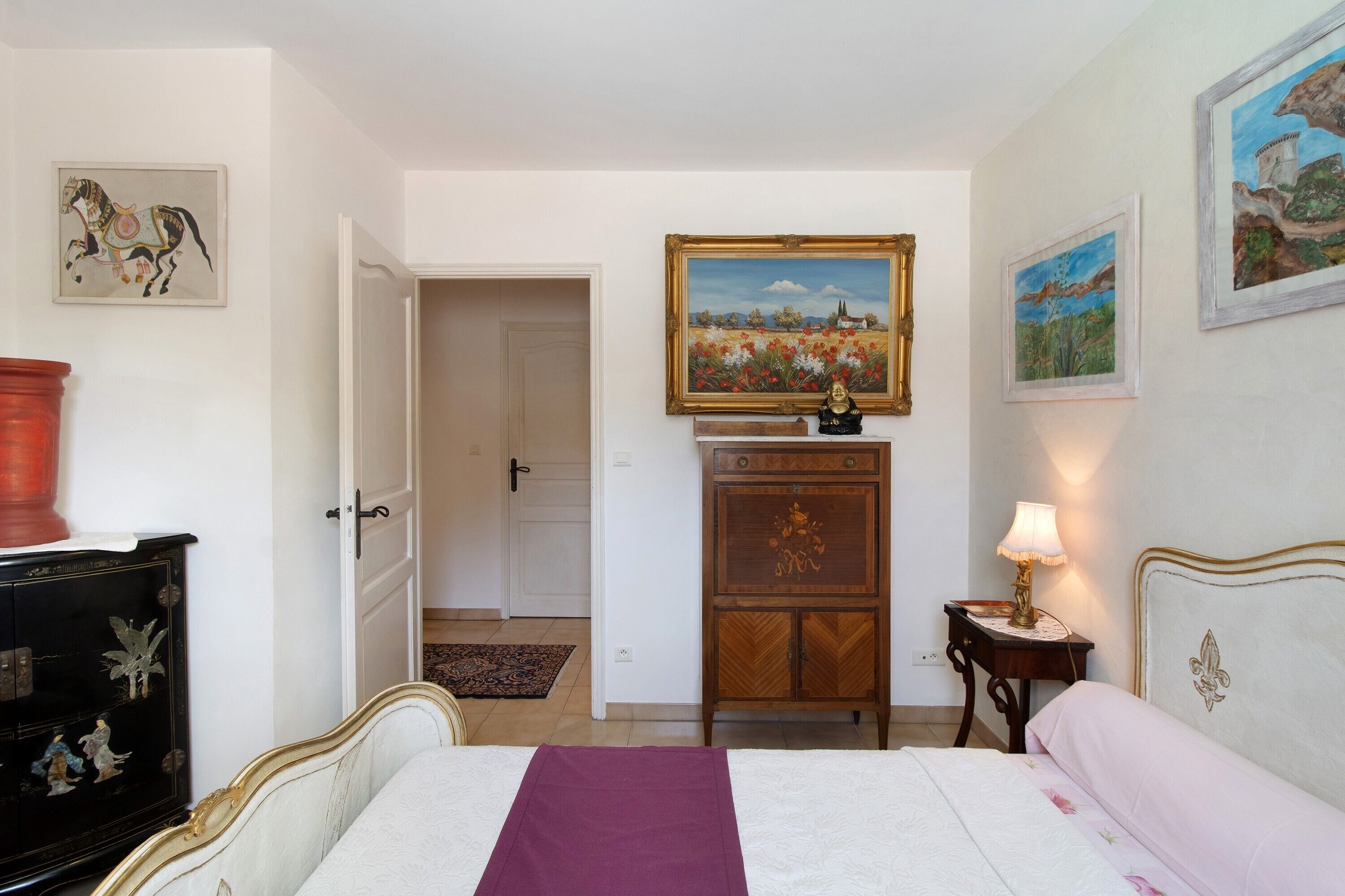 Een slaapkamer met een bed, nachtkastje, dressoir en een deur die naar een andere kamer leidt.