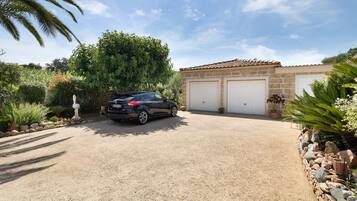 Uma entrada de cascalho leva a uma garagem para dois carros com um edifício de pedra, tudo cercado por vegetação e uma palmeira.