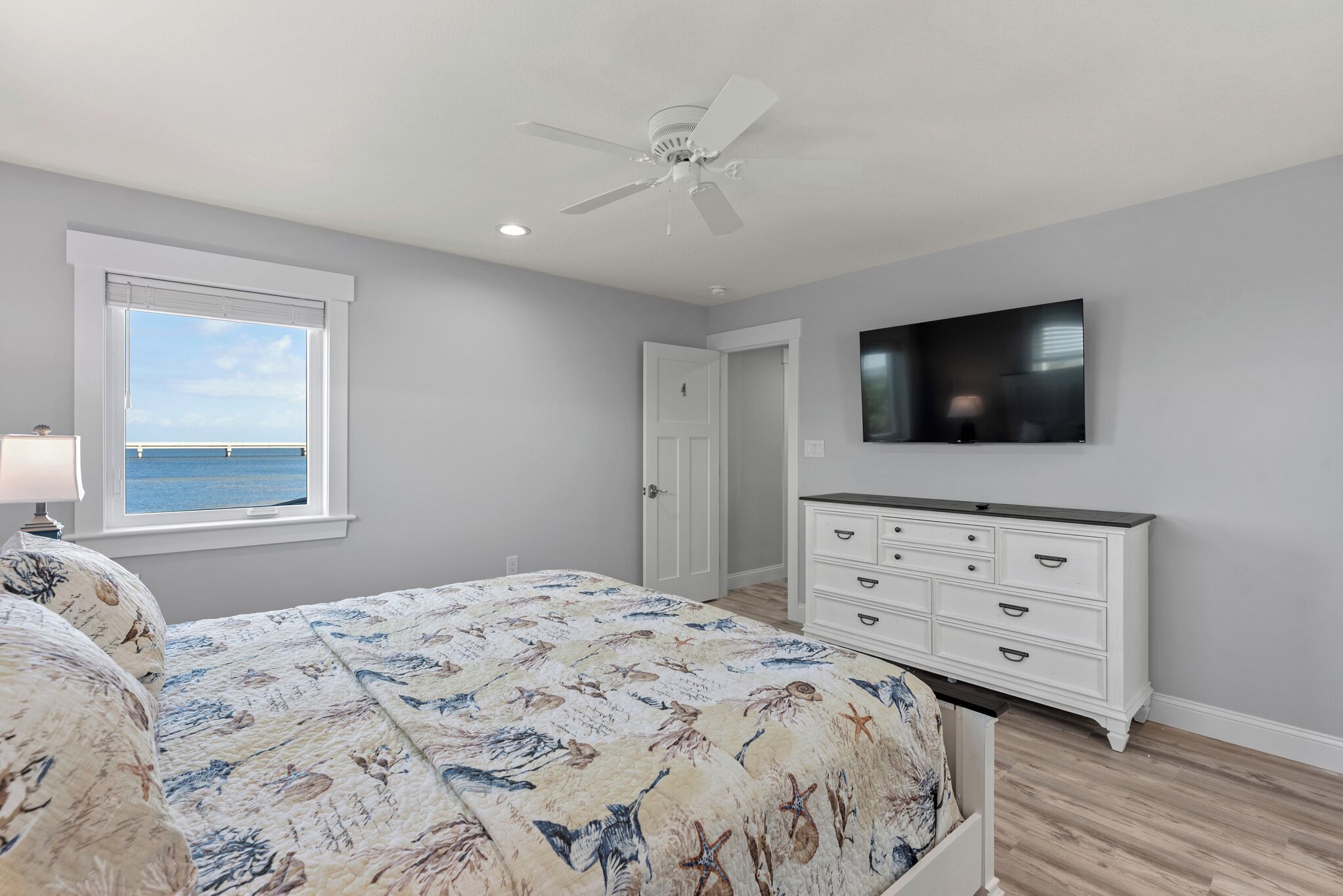 A bedroom with a bed, a dresser, a ceiling fan, a TV, and a window with a view of the ocean.