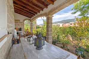 Une terrasse couverte avec des colonnes en pierre, un plafond en bois rustique et une vue sur un jardin agrémenté de plantes en pot et d’une chaîne de montagnes lointaine.