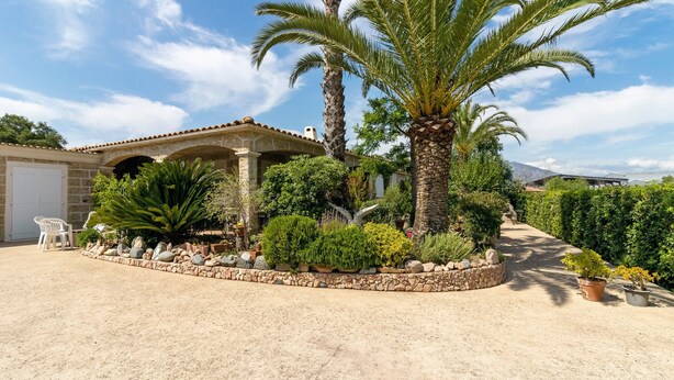 A house with a covered patio, palm trees, and a garden with various plants and a stone wall. - Bed and Breakfast 'Gautama' with Mountain View, Shared Terrace and Shared Garden (SARROLA CARCOPINO)