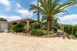 A house with a covered patio, palm trees, and a garden with various plants and a stone wall.