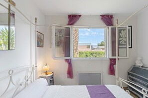 Een slaapkamer met een bed, een nachtkastje met lamp, een dressoir en een raam met gordijnen.