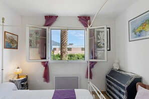 Een slaapkamer met een bed, een nachtkastje met een lamp, een dressoir, een raam met gordijnen en uitzicht op gebouwen en groen.