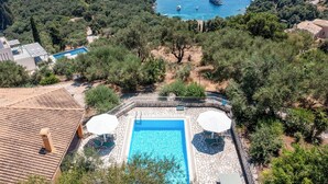 Zona de piscina con vista despejada al mar y barcos, rodeada de árboles y un muro de piedra.