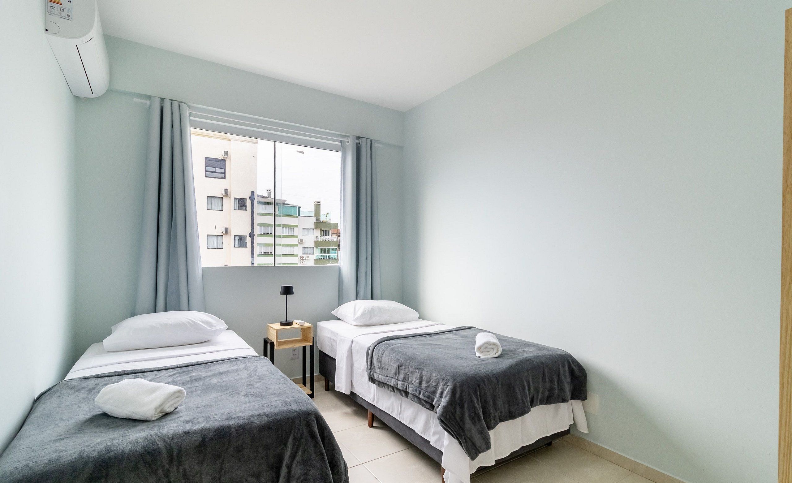 A hotel room with two single beds, a small bedside table with a lamp, a window with curtains, and a view of buildings outside.