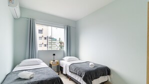 A hotel room with two single beds, a small bedside table with a lamp, a window with curtains, and a view of buildings outside.