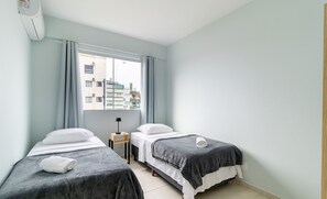 Ein Hotelzimmer mit zwei Einzelbetten, einem kleinen Nachttisch mit Lampe, einem Fenster mit Vorhängen und Blick auf Gebäude.