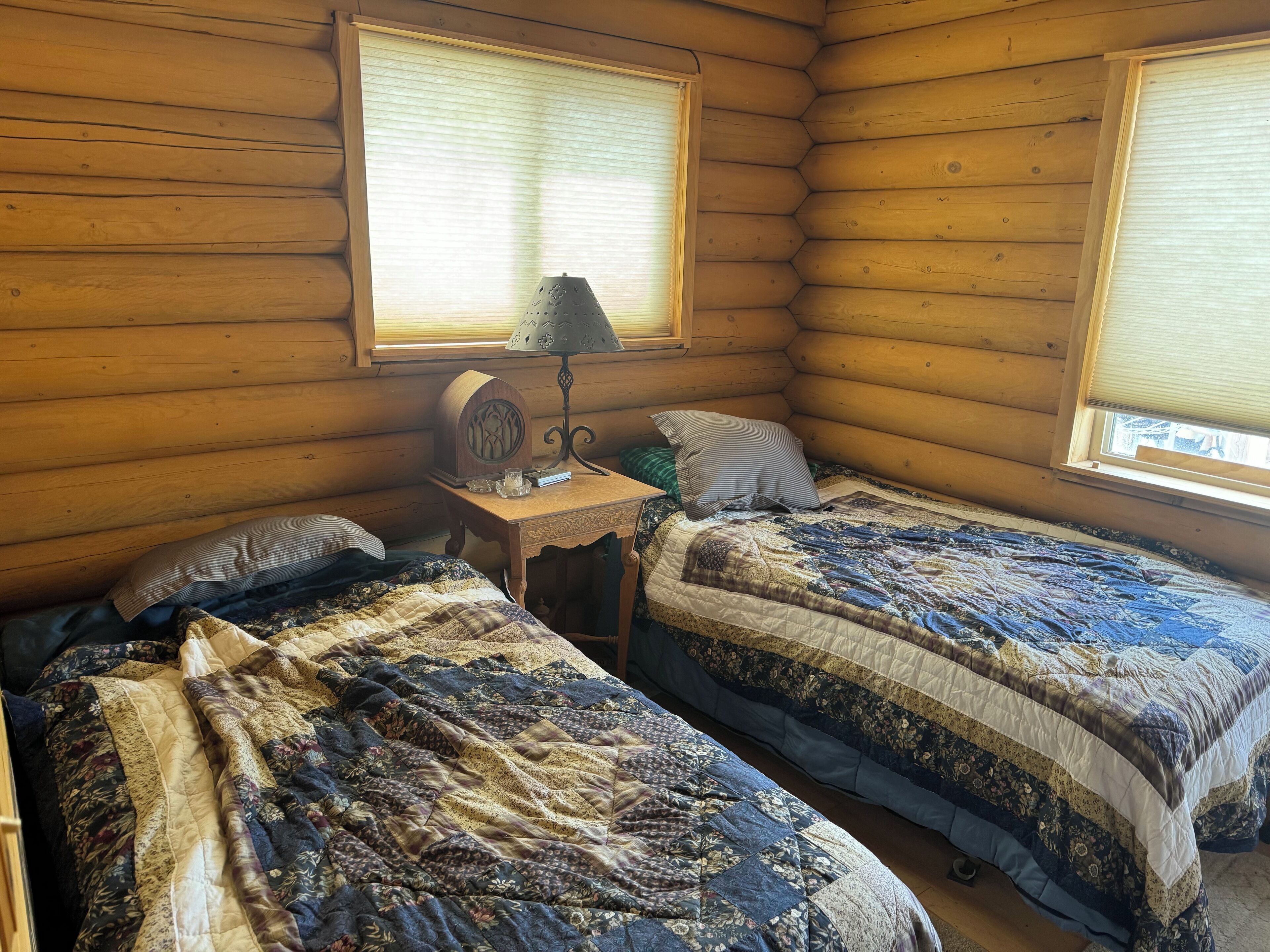 Una habitación tipo cabaña con dos camas, un cabecero de madera, una mesita con lámpara y una ventana con persianas.