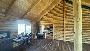 Interieur van een blokhut met een houten vloer, een werkbank, een raam en een zichtbare keuken door een open deur.