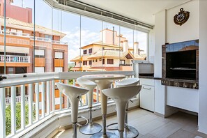 A balcony with white bar stools, a built-in grill, and a view of neighboring buildings. - Apto no Open Shopping 600m da praia IBI307 (Florianópolis)