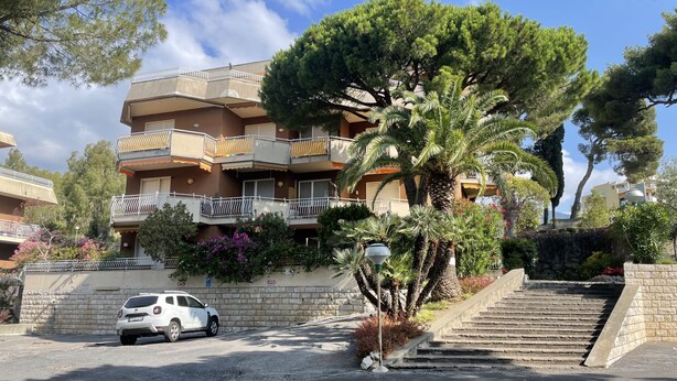 A multi-story residential building with balconies, a car parked in front, and a staircase leading to the entrance.