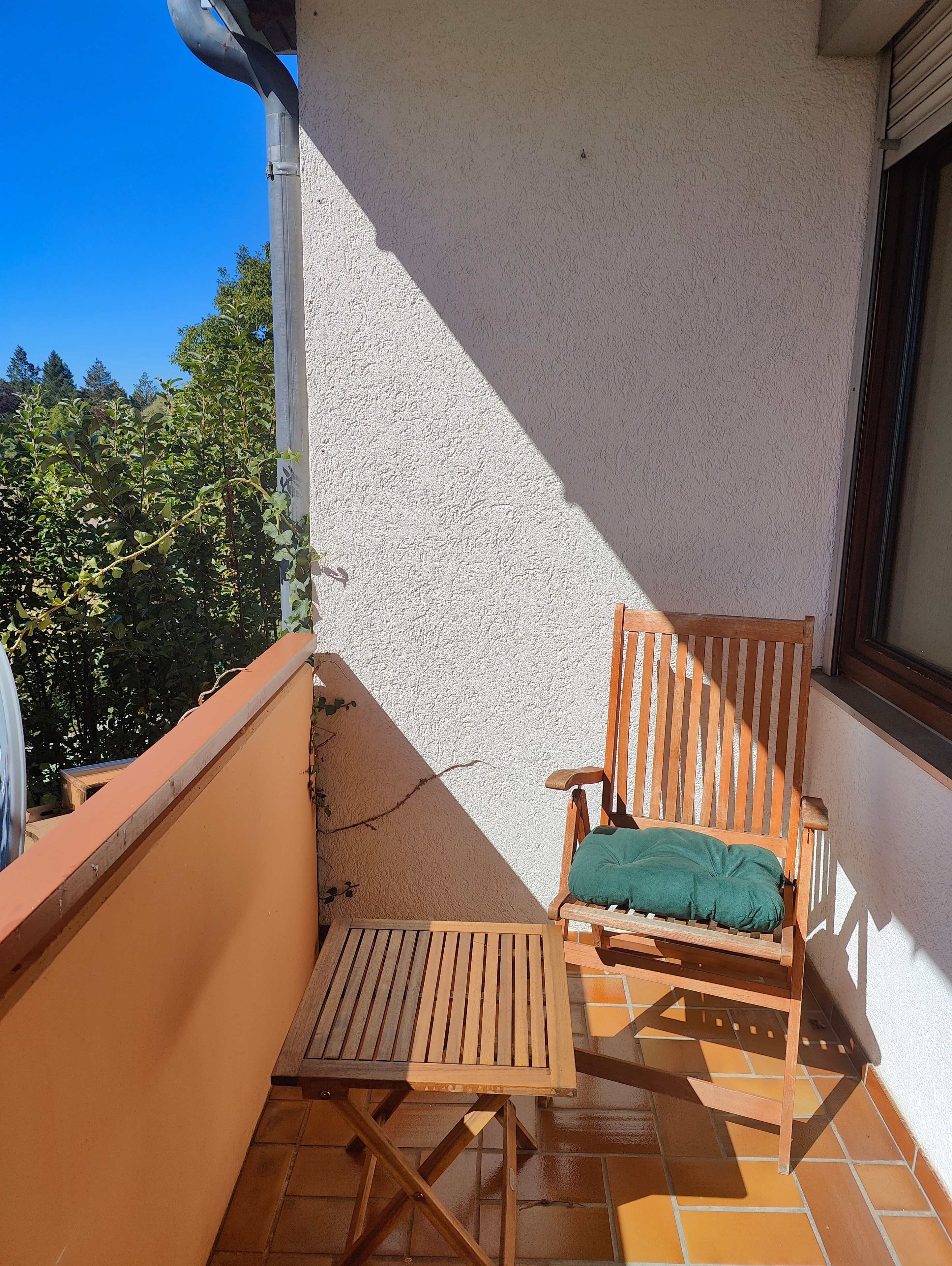 Un balcón con una silla y mesa de madera, una pared blanca con textura y vistas a árboles.