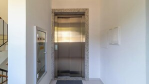 Um elevador em um corredor com porta de vidro, estrutura metálica, tapete de entrada e um interruptor de luz fixado na parede.