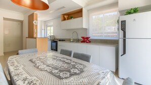 Uma cozinha moderna com armários brancos, forno embutido e geladeira. Há uma área de jantar com toalha de mesa estampada e cadeiras.