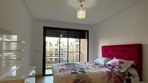 A bedroom with a bed, a dresser, a nightstand, a chandelier, and a sliding glass door to a balcony. - Prestigia Luxury apartment near the beach "Plage Des Nations" Sale/Rabat/Airport (Rabat-Salé-Kénitra)