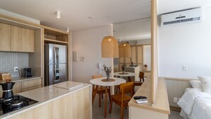 Una cocina moderna con armarios de madera, un refrigerador empotrado y una cafetera. Hay un área de comedor con una mesa redonda y sillas, y una cama con ropa blanca en la habitación contigua.
