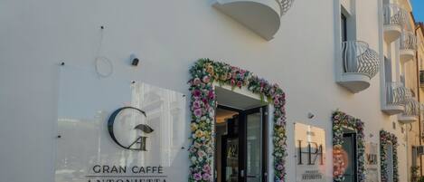 Fachada de hotel con entrada despejada, un letrero que dice GRAN CAFFÈ ANTONIETTA y arreglos florales decorativos.