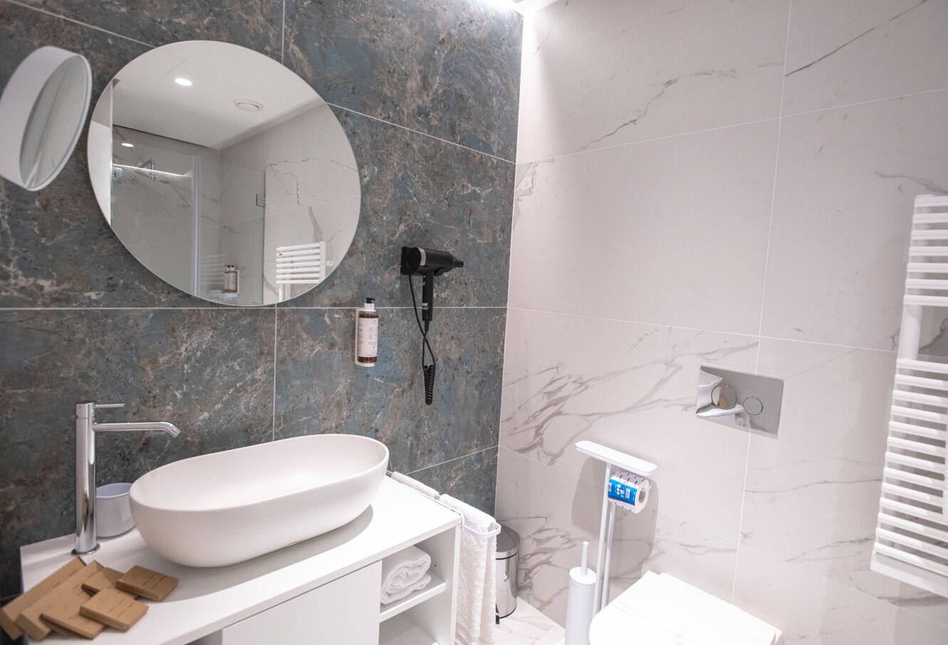 A modern bathroom with a large oval sink, a round mirror, and a handheld hair dryer mounted on the wall.