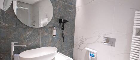 A modern bathroom with a large oval sink, a round mirror, and a handheld hair dryer mounted on the wall.