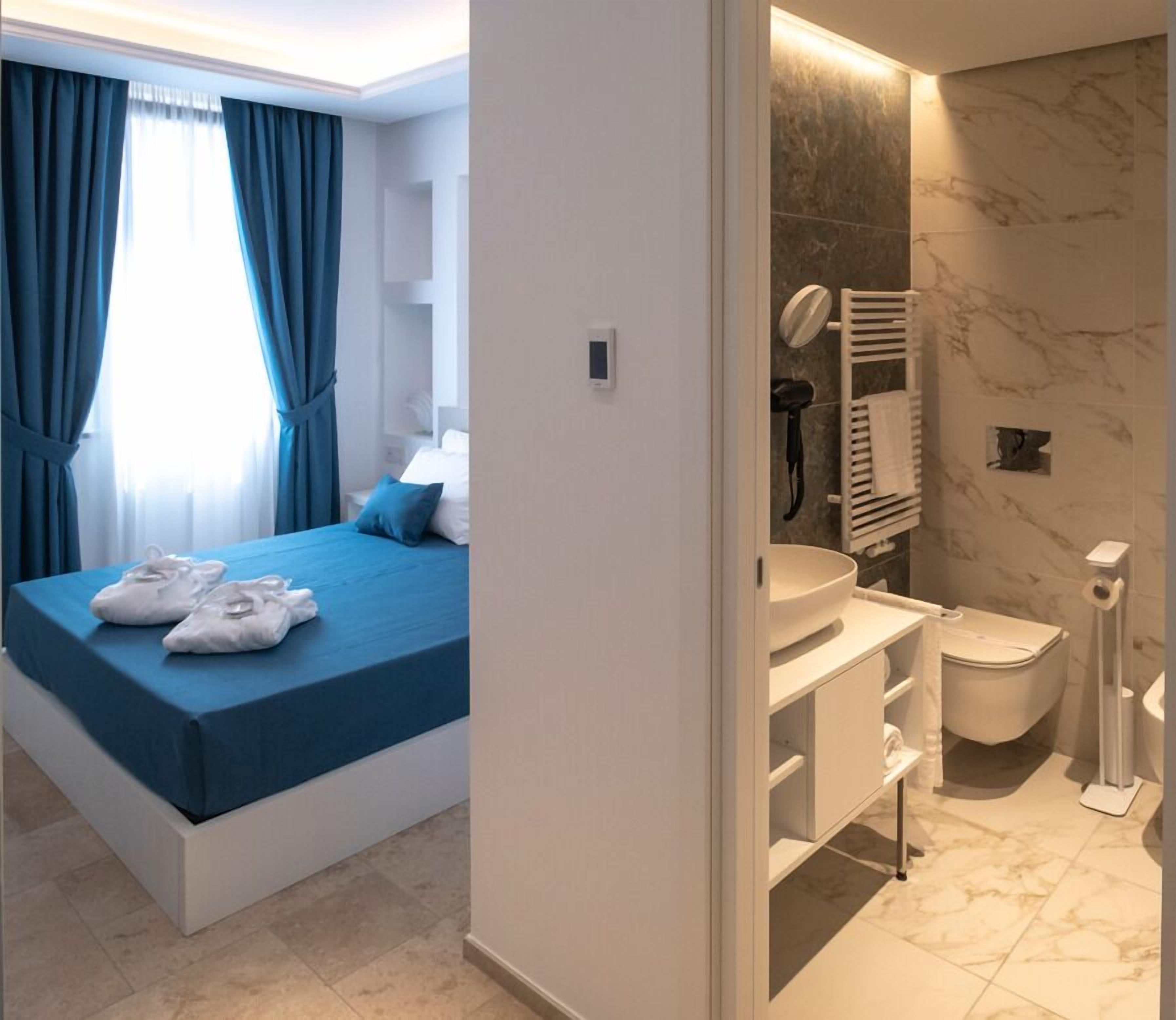 A modern bedroom with a bed, a nightstand, and a window with curtains. Adjacent is a bathroom with a sink, mirror, and toilet.