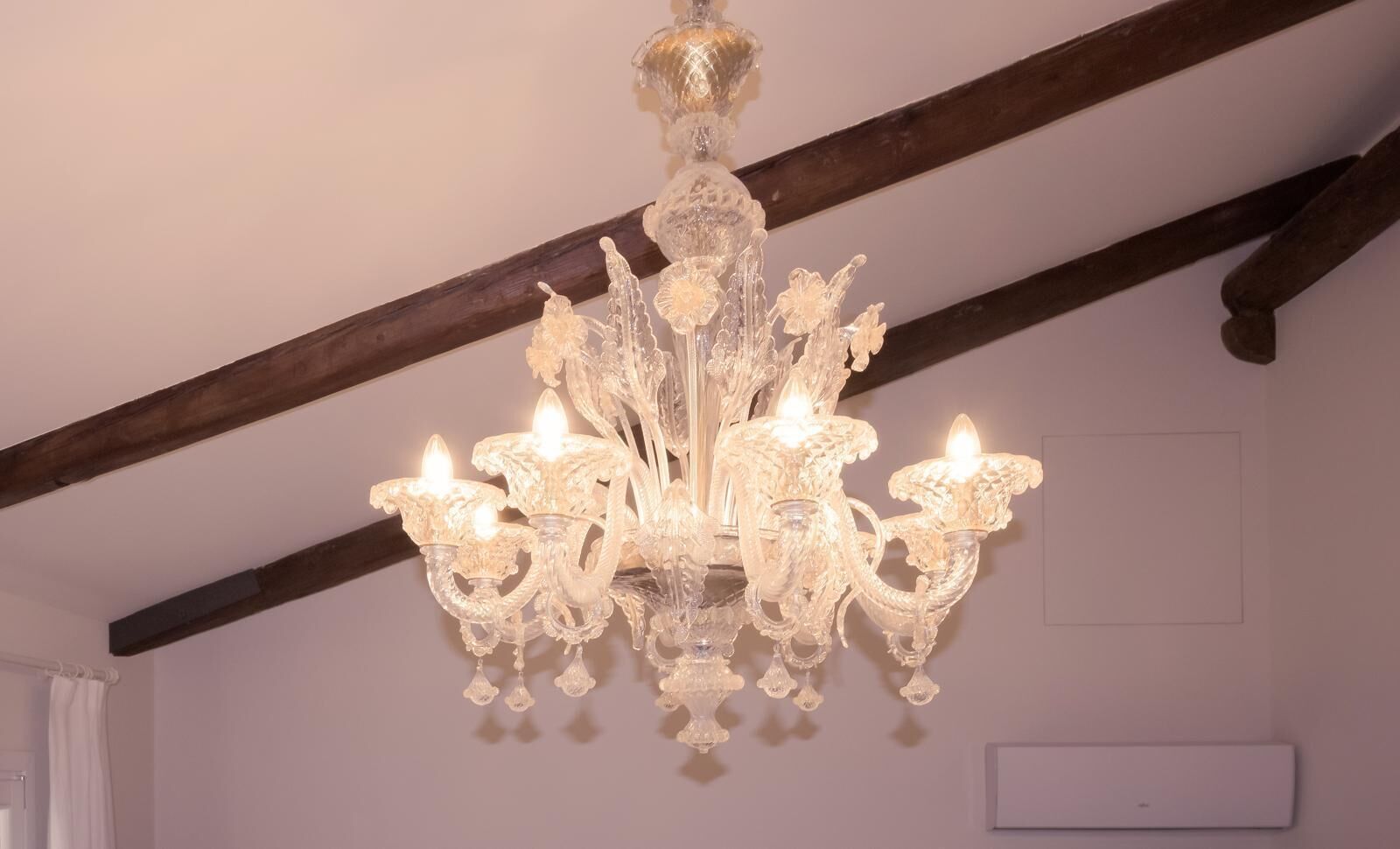Un lustre orné de plusieurs lampes en forme de bougies, une poutre en bois au plafond et un mur blanc avec un tableau de distribution électrique vide.