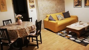 A living room with a yellow sofa, a round table with chairs, and a coffee table. There are framed pictures on the wall and a vase with flowers on the table.