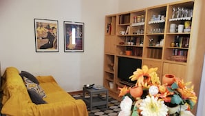 A living room with a sofa, a coffee table with a vase of flowers, a television, and a bookshelf filled with books and decorative items.