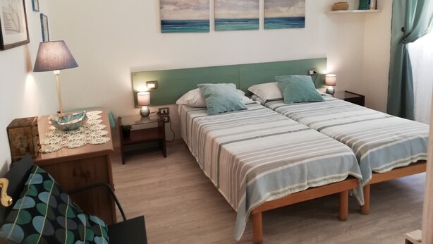 A double bed with striped bedding, two bedside tables with lamps, a small wooden nightstand with a lamp, a framed picture on the wall, and a shelf with books and a vase.