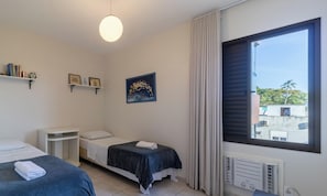 A bedroom with two beds, a bedside table, a wall-mounted shelf with books and a clock, a painting, a window with curtains, and a radiator.