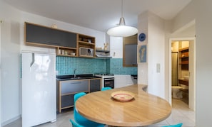 A modern kitchen with a white refrigerator, a stove, and a sink. There is a wooden dining table with chairs and a hanging light fixture.