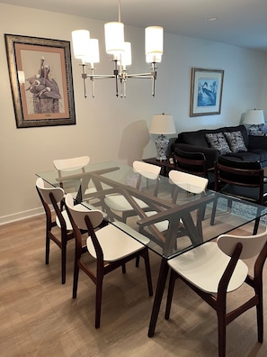 A dining area with a glass table and chairs, a chandelier, framed artwork, and a sofa with cushions.