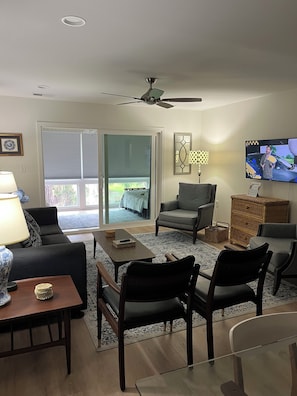 A living room with a sofa, armchairs, a coffee table, a TV, a ceiling fan, and a view through a sliding door to a balcony.
