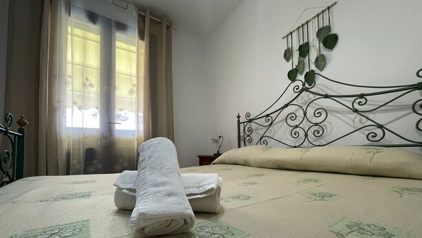 A neatly made bed with a floral-patterned bedspread, a decorative wall hanging with leaves, and a window with sheer curtains.