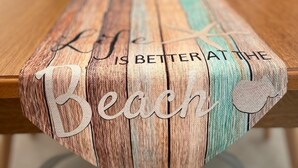A wooden table with a woven table runner featuring the text "Life is better at the beach."