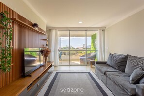 A modern living room with a grey sofa, a flat-screen TV on a wooden console, a large sliding glass door leading to an outdoor balcony, and a decorative plant on the left. - Full Apartment at Ingleses Beach-sc Gge203 (Florianópolis)