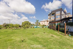 A grassy outdoor area with a playground and a multi-story building in the background. - Full Apartment at Ingleses Beach-sc Gge203 (Florianópolis)