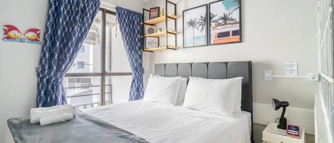 Una habitación de hotel con una cama, una mesita de noche con lámpara y una ventana con cortinas azules.