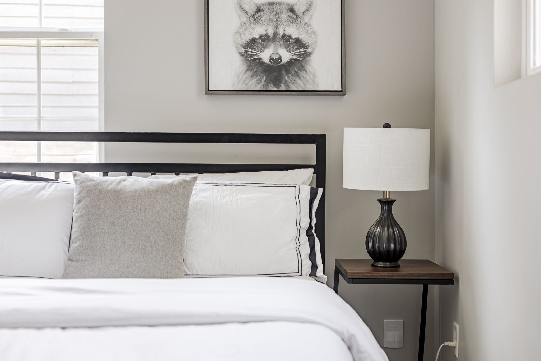 Un lit bien fait, avec une literie blanche et un oreiller gris, une tête de lit à cadre noir, une table de chevet avec une lampe, et une œuvre d’art représentant un raton laveur, encadrée, accrochée au mur.