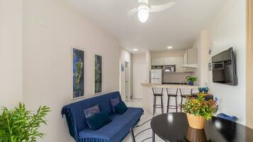 A modern living room with a sofa, a dining table, and chairs. There is a kitchen area with a microwave and a TV.