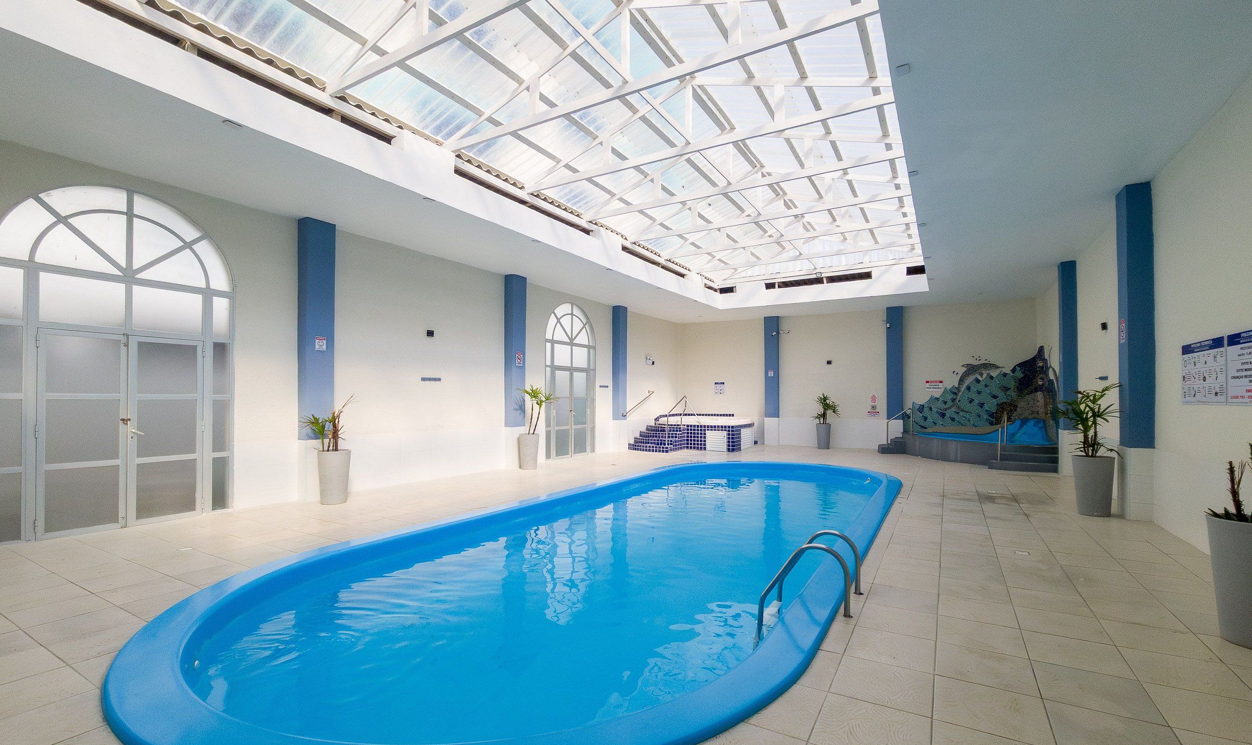 An indoor swimming pool with a skylight ceiling, a hot tub, and potted plants.