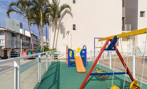 Un parque infantil con toboganes y estructuras para escalar, rodeado por una valla, palmeras y edificios residenciales.