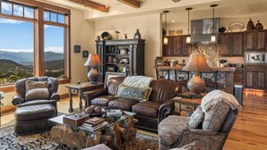 A spacious living room with leather furniture, a large window offering a mountain view, and a modern kitchen in the background. - Cowboy Lux - Moonlight Basin Ski In/Out (Big Sky)