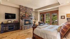 A bedroom with a stone fireplace, a large bed, a TV, and a view of greenery outside. - Cowboy Lux - Moonlight Basin Ski In/Out (Big Sky)