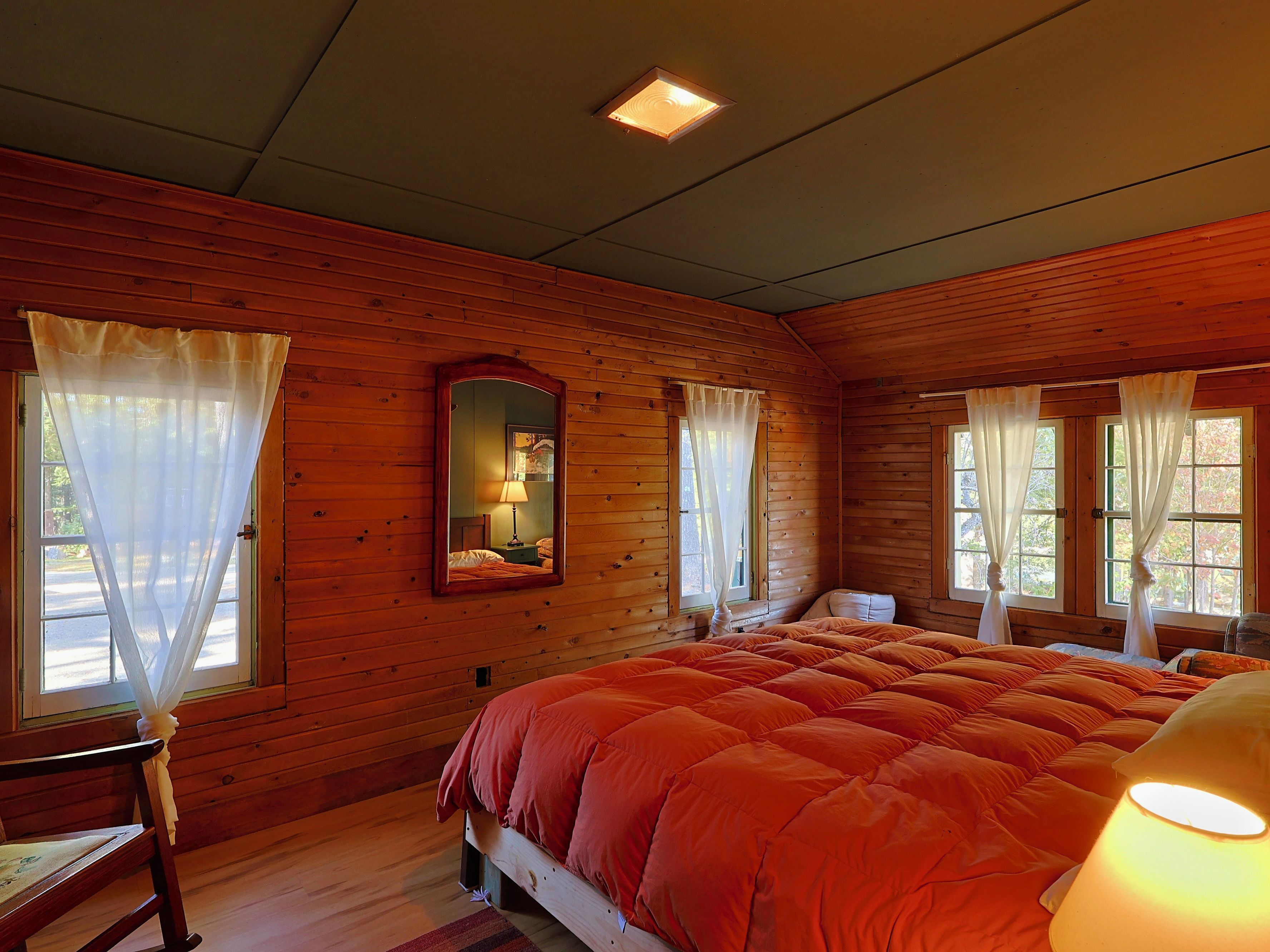 A wooden cabin room with a bed, a chair, a table, a mirror, and windows with curtains.