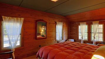 A wooden cabin room with a bed, a chair, a table, a mirror, and windows with curtains.